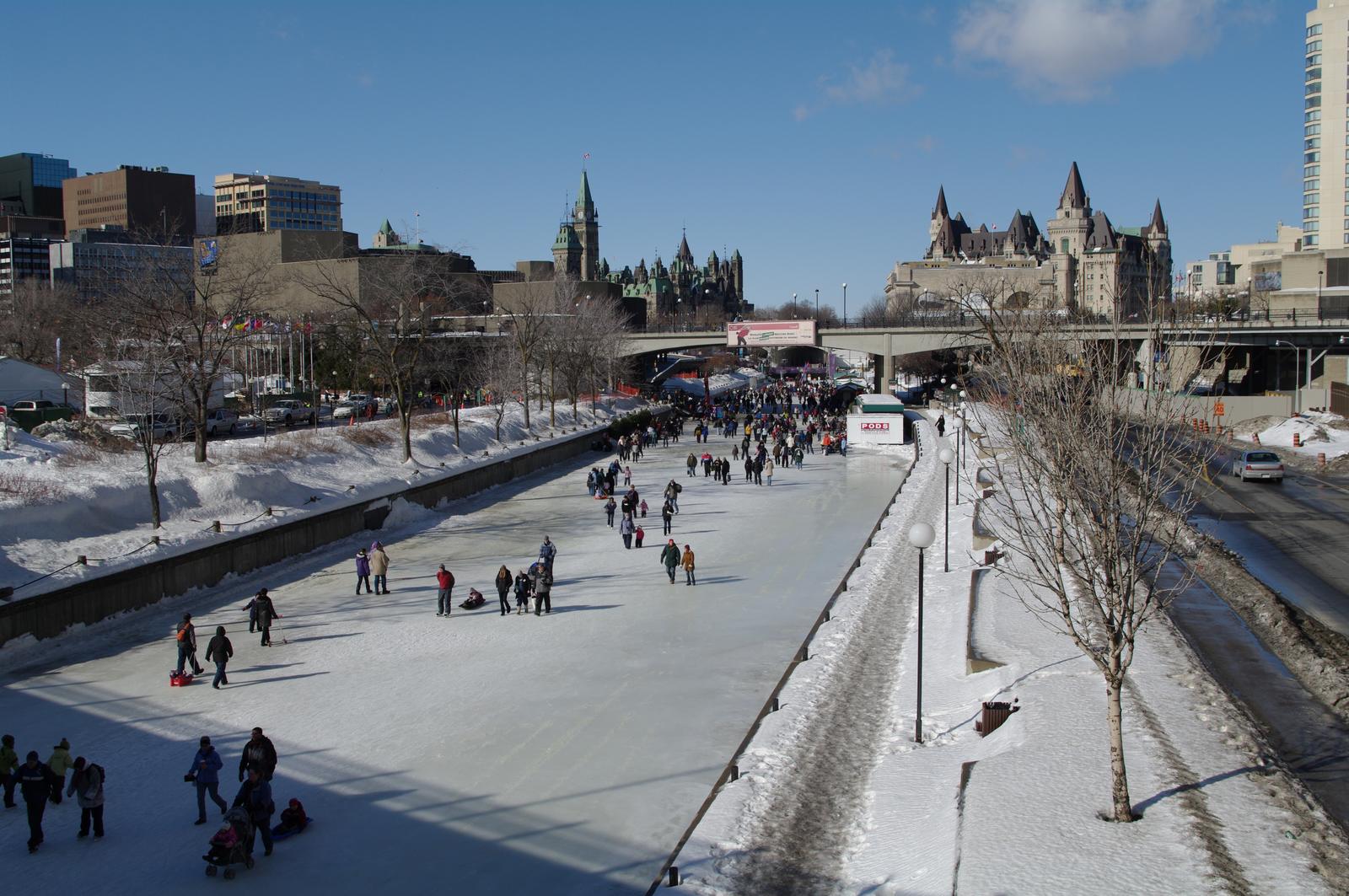Skaters on the frozen Rideau Canal Skateway in downtown Ottawa on a sunny winter day, with the Château Laurier and the Parliament buildings in the background.